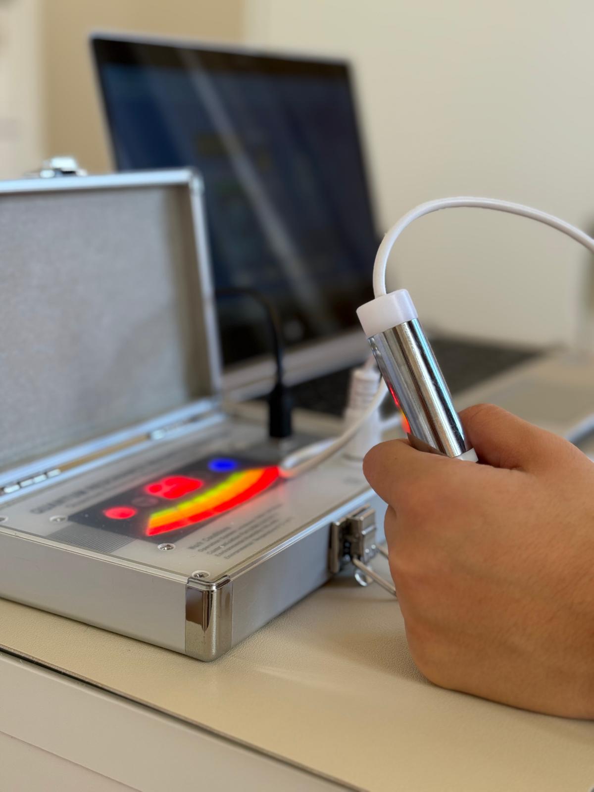 Kleiner Metallkoffer mit einem Metallrohr für den Vitaltest im OndaZentrum: Hand, die einen an Kabel angeschlossenen Metallstab hält, im Hintergrund Metallkoffer mit verschiedenen Lichtern